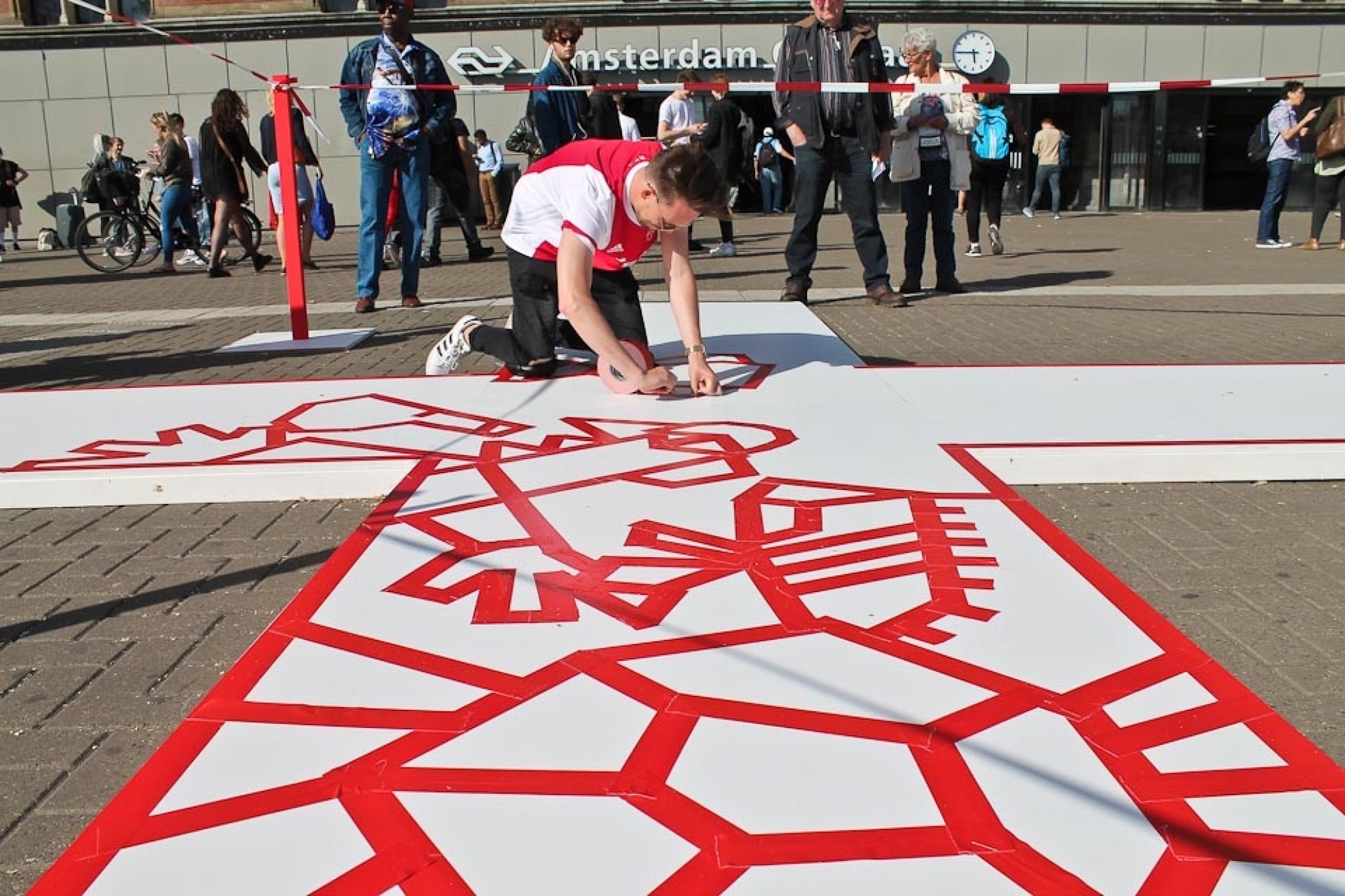 Art piece created by Pablo Lücker with red tape in front of Amsterdam Central Station for Adidas X Ajax UEFA Finals. title=